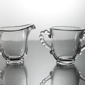 K&ouml;nnte beinhalten: Ein Set aus zwei durchsichtigen Glaskr&uuml;gen f&uuml;r Sahne und Zucker mit perlenbesetzten Griffen.