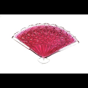 May include: A decorative, ruby-red glass fan-shaped dish. The fan features a detailed, textured pattern and a scalloped edge. The base is clear glass. The dish is designed to be a decorative piece.