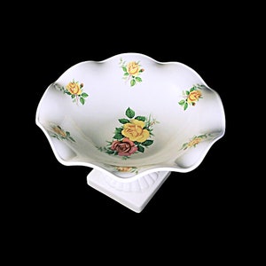 Puede incluir: Tazón de cerámica blanca con borde festoneado y diseño floral con rosas amarillas, rosas y rojas. El tazón está sobre una base de pedestal.
