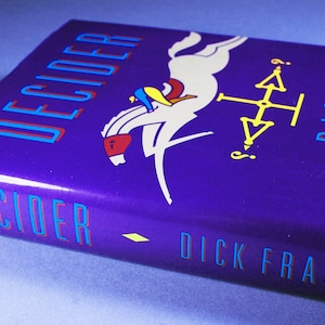 May include: A purple hardcover book with the title "DECIDER" in blue and red lettering. The book cover features a white horse jumping over a yellow compass with a question mark. The author's name, "DICK FRANCIS", is printed in blue and red lettering at the bottom of the cover.