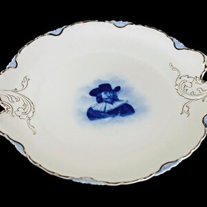 May include: A white porcelain serving platter with gold trim and a blue and white image of a man in a hat and ruff. The platter has two ornate handles.