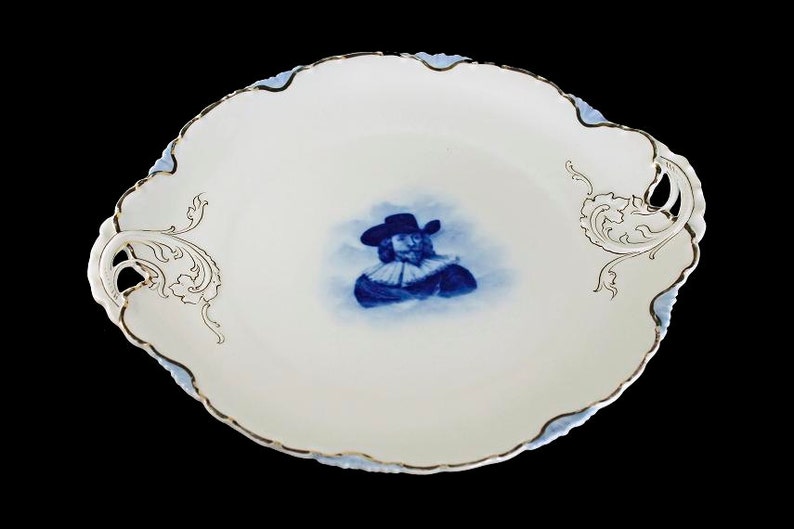 May include: A white porcelain serving platter with gold trim and a blue and white image of a man in a hat and ruff. The platter has two ornate handles.