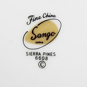 Op de afbeelding: Fine China Sango Japan Sierra Pines 6608 C. Een wit keramiek bord met een zwart en gouden logo.