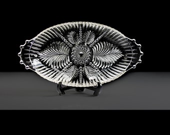 May include: A clear glass serving dish with a decorative pattern of leaves and a beaded center. The dish is oval shaped and has a raised edge.