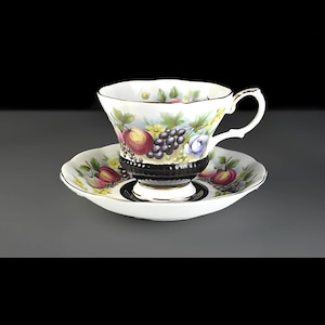 以下が含まれることがあります： ヴィンテージのティーカップとソーサーのセット。白い磁器のカップとソーサーには、カラフルな果物と花の模様が描かれています。カップには黒いバンドと繊細なハンドルが付いています。ソーサーはカップのデザインを反映しています。