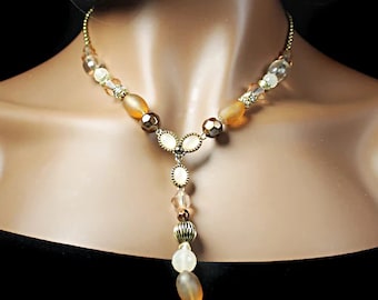 Op de afbeelding: Een gouden en bruine kralenketting met een Y-vormig ontwerp en een lange, hangende hanger. De ketting heeft verschillende kralen, waaronder ovale, ronde en gefacetteerde kralen.
