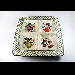 Puede incluir: Una bandeja de servir de cerámica blanca con cuatro compartimentos, cada uno con un diseño de fruta diferente. La bandeja tiene un borde de rejilla verde y un acabado desgastado.