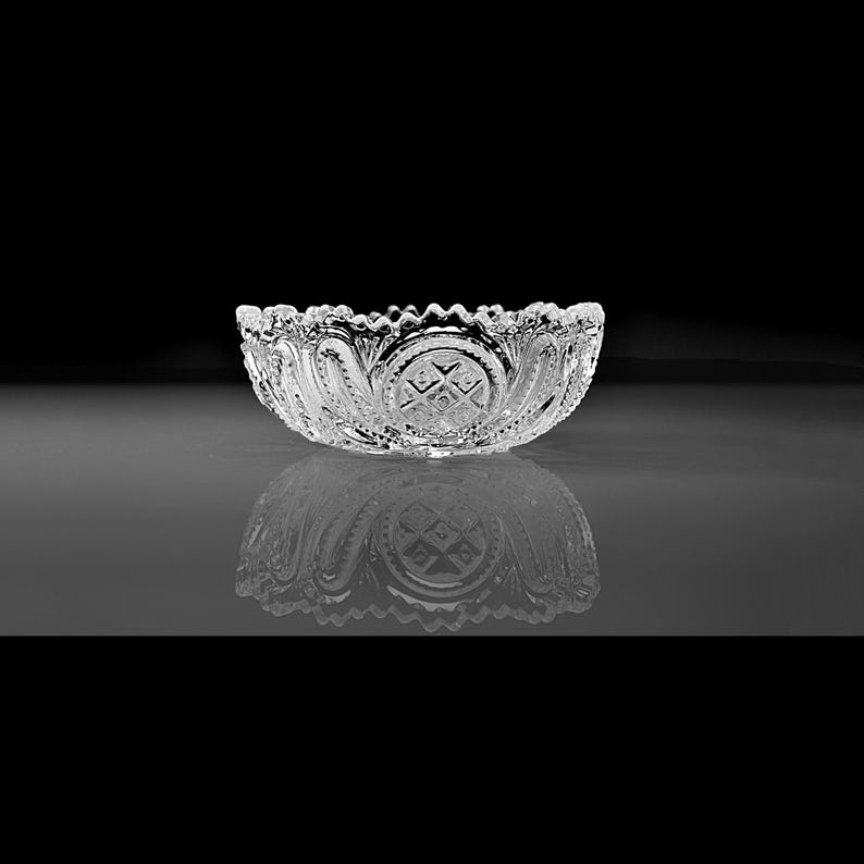 Pu&ograve; includere: Una ciotola di vetro trasparente e decorativa con un bordo smerlato e intricati motivi incisi. Il design della ciotola include motivi floreali e geometrici. La ciotola &egrave; esposta su una superficie riflettente su sfondo nero.