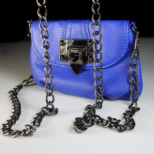 Pode incluir: Uma pequena bolsa azul cobalto com uma superf&iacute;cie texturizada e uma al&ccedil;a de corrente de metal. A bolsa apresenta um fecho retangular e uma aba. A corrente &eacute; drapeada na frente da bolsa.