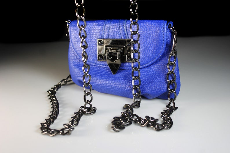 Pode incluir: Uma pequena bolsa azul cobalto com uma superf&iacute;cie texturizada e uma al&ccedil;a de corrente de metal. A bolsa apresenta um fecho retangular e uma aba. A corrente &eacute; drapeada na frente da bolsa.