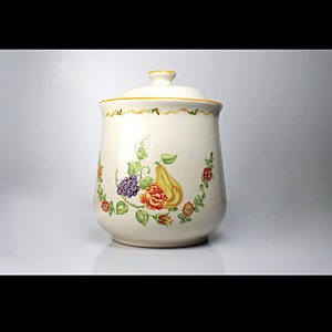 May include: A white ceramic canister with a lid, featuring a floral design with a pear, grapes, and a rose. The canister has a yellow trim around the lid and base.