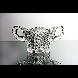 May include: A clear glass sugar bowl with two handles and a cut glass design. The bowl is reflecting on a shiny surface.