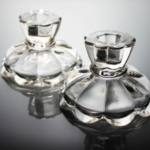 Puede incluir: Dos candelabros de vidrio transparente con borde festoneado y dise&ntilde;o acanalado. Los candelabros est&aacute;n sobre una superficie reflectante.