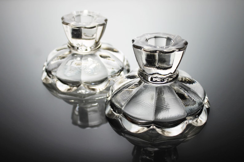 Puede incluir: Dos candelabros de vidrio transparente con borde festoneado y dise&ntilde;o acanalado. Los candelabros est&aacute;n sobre una superficie reflectante.
