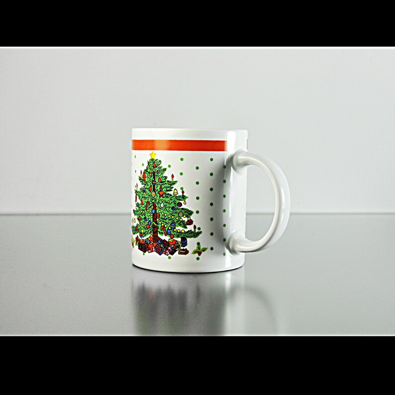 May include: White ceramic mug featuring a red and green Christmas tree design. The tree is adorned with baubles and presents. The mug has a white handle.