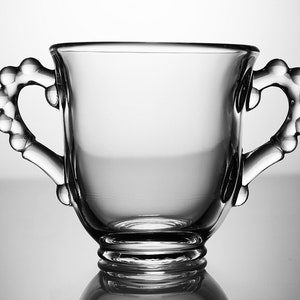 K&ouml;nnte beinhalten: Eine klare Glas Tasse mit zwei perlenbesetzten Griffen. Die Tasse hat einen abgerundeten Boden und eine breite, offene Oberseite.