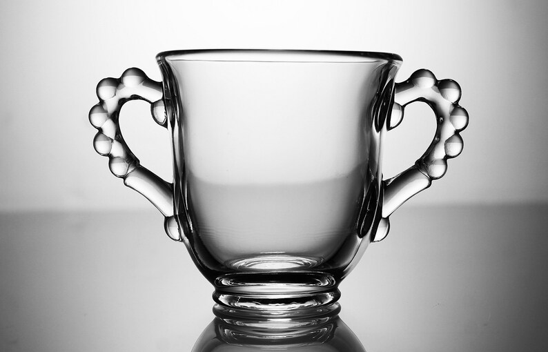 K&ouml;nnte beinhalten: Eine klare Glas Tasse mit zwei perlenbesetzten Griffen. Die Tasse hat einen abgerundeten Boden und eine breite, offene Oberseite.