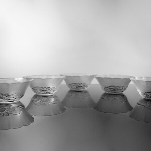May include: Five clear glass bowls with a scalloped rim and floral design on the base. The bowls are arranged in a row, reflecting on a glossy surface. The image is in black and white.
