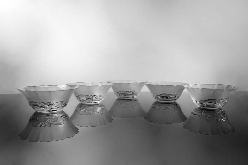 May include: Five clear glass bowls with a scalloped rim and floral design on the base. The bowls are arranged in a row, reflecting on a glossy surface. The image is in black and white.