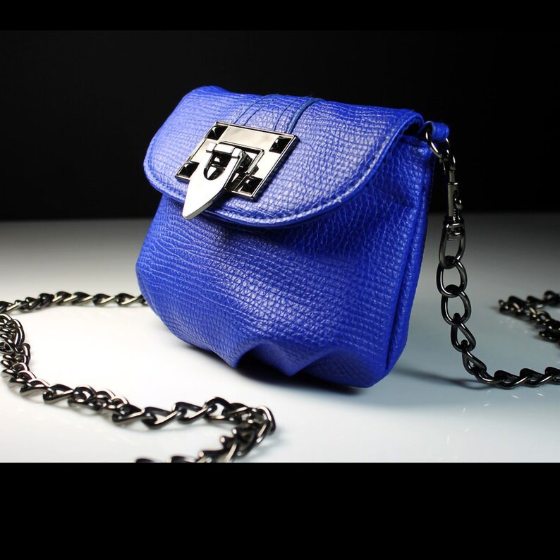 Pode incluir: Uma pequena bolsa azul cobalto com um acabamento texturizado. A bolsa apresenta um fecho prateado e uma al&ccedil;a de corrente preta. A bolsa tem uma forma arredondada com um fecho de aba.