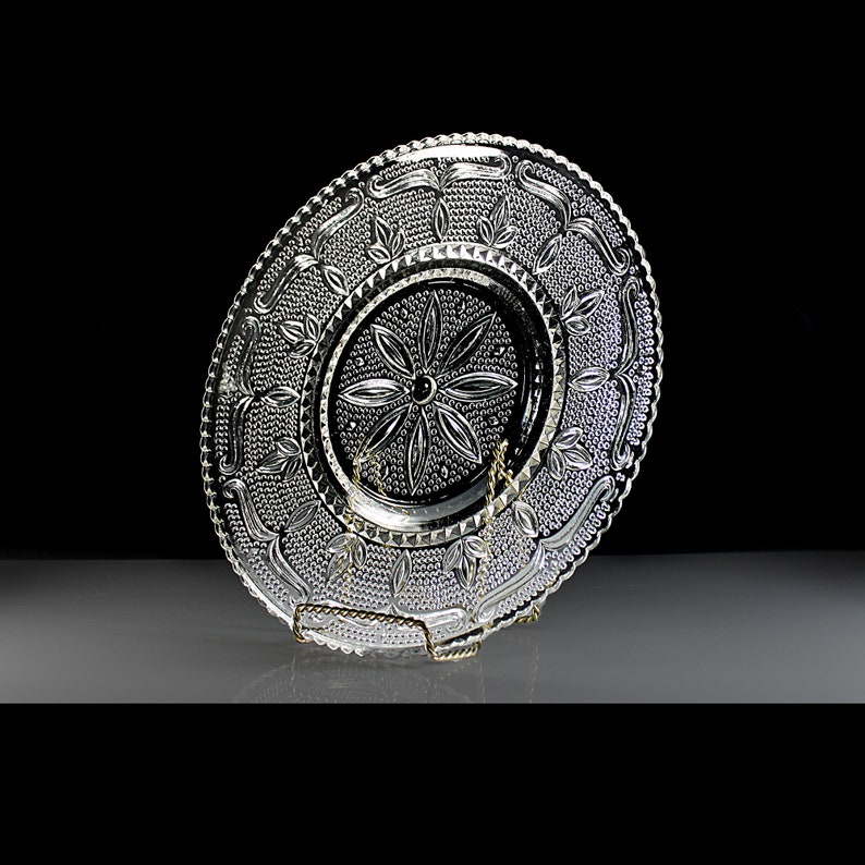 Op de afbeelding: Een heldere glazen bord met een decoratief, reli&euml;fpatroon van bladeren en bloemen. Het bord heeft een gegolfde rand en staat op een standaard.
