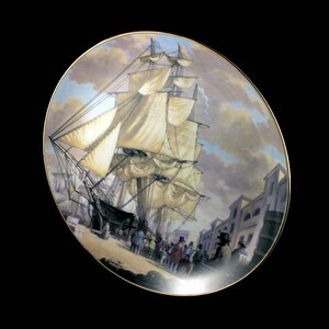 May include: Decorative plate featuring a detailed painting of a tall ship with billowing sails docked near a town. The plate has a gold rim and a classic, historical theme. The artwork depicts a bustling scene with figures on the shore.