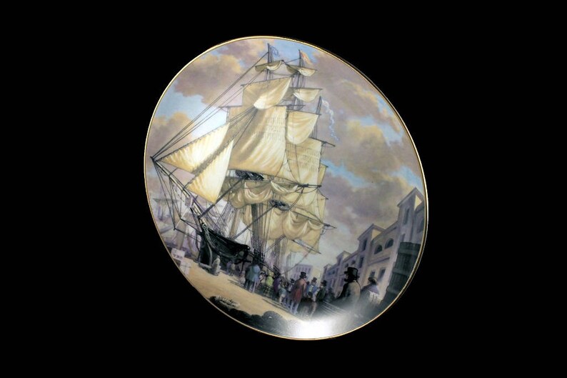May include: Decorative plate featuring a detailed painting of a tall ship with billowing sails docked near a town. The plate has a gold rim and a classic, historical theme. The artwork depicts a bustling scene with figures on the shore.