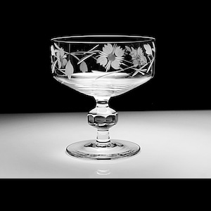 Könnte beinhalten: Eine klare Champagner-Coupe aus Glas mit einem zarten, geätzten Blumenmuster im Kelch. Die Coupe hat einen kurzen Stiel und einen breiten Standfuß.