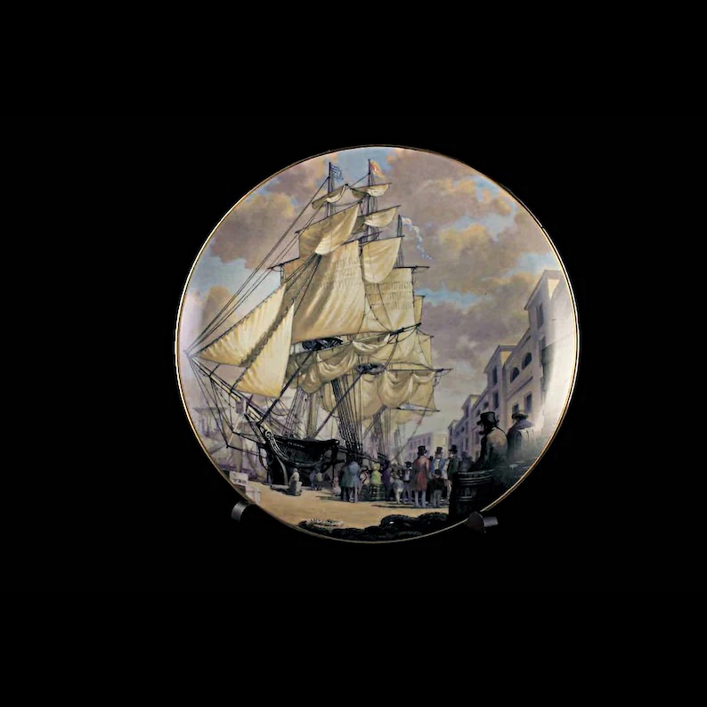May include: Decorative plate featuring a detailed painting of a tall ship docked at a port, with figures on the pier and buildings in the background. The plate has a gold rim and is displayed on a black stand.