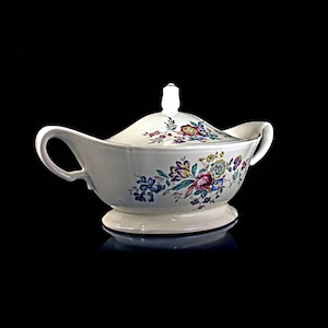 Puede incluir: Un plato de servir de porcelana blanca con tapa, que presenta un diseño floral en tonos de rosa, morado, amarillo y verde. El plato tiene una forma redondeada con dos asas y una base elevada.
