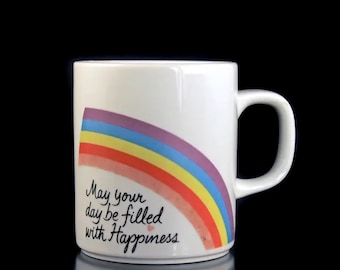 Op de afbeelding: Een witte keramische mok met een regenboogontwerp en de tekst "May your day be filled with Happiness".