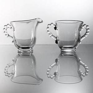 K&ouml;nnte beinhalten: Ein Set aus zwei durchsichtigen Glaskr&uuml;gen f&uuml;r Sahne und Zucker mit perlenbesetzten Griffen. Die Kr&uuml;ge sind leer und stehen auf einer reflektierenden Oberfl&auml;che.