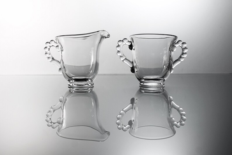 K&ouml;nnte beinhalten: Ein Set aus zwei durchsichtigen Glaskr&uuml;gen f&uuml;r Sahne und Zucker mit perlenbesetzten Griffen. Die Kr&uuml;ge sind leer und stehen auf einer reflektierenden Oberfl&auml;che.