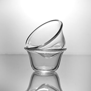 May include: Two clear glass bowls stacked on top of each other. The top bowl is tilted, revealing its interior. The bowls have a rounded shape with a slightly flared rim. The image is in black and white.