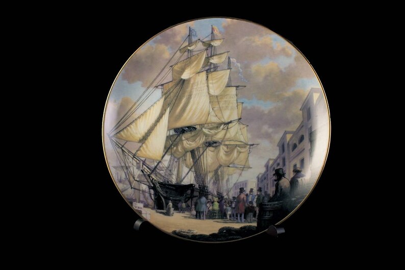 May include: Decorative plate featuring a detailed painting of a tall ship docked at a port. The artwork depicts the ship with billowing sails, figures on the dock, and buildings in the background. The plate has a gold rim.