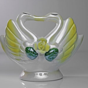 May include: White ceramic swan figurine with yellow wings and a green and yellow rose in the center. The swans are facing each other with their necks forming a heart shape.