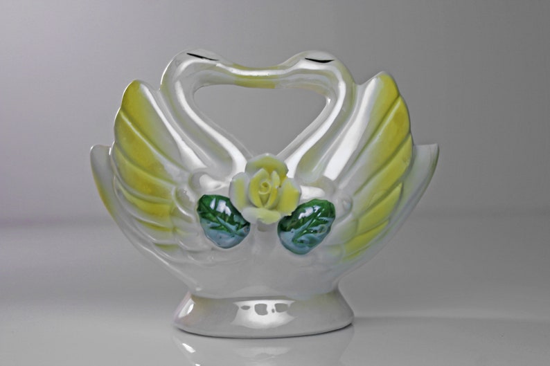 May include: White ceramic swan figurine with yellow wings and a green and yellow rose in the center. The swans are facing each other with their necks forming a heart shape.