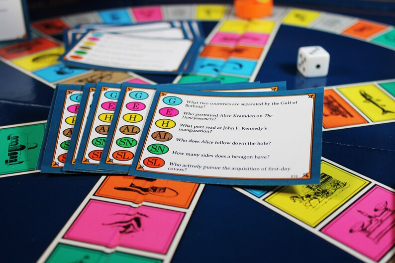 以下が含まれることがあります： 青いゲームボードにカラフルなマスと、トリビア問題が書かれた青いカードの山があります。カードには、文字が書かれた異なる色の円があります。カードのテキストは、「ボトニア湾で隔てられている2つの国は？」、「The Honeymoonersでアリス・クラムデンを演じたのは誰？」、「ジョン・F・ケネディの就任式で詩を読んだのは誰？」、「アリスは穴に誰を追いかけていくのですか？」、「六角形はいくつ辺がありますか？」、「初日カバーの取得を積極的に追求するのは誰ですか？」です。