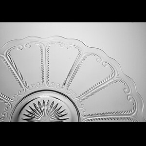 Pu&ograve; includere: Un piatto da portata in vetro trasparente con un bordo ondulato e un motivo decorativo di volute e linee ripetute.