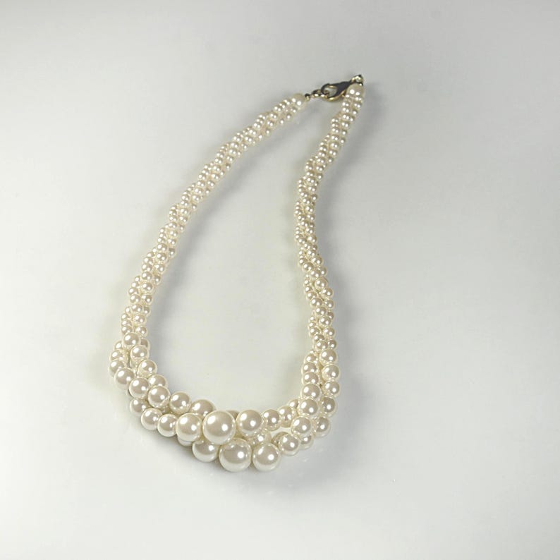 May include: A cream pearl necklace with a multi-strand design. The necklace has a clasp and a graduated arrangement of pearls, with larger pearls at the centre. The pearls are round and have a lustrous finish.