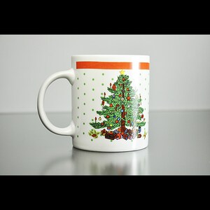 May include: White ceramic mug with a red and orange stripe around the top. The mug features a green Christmas tree with red and gold baubles, a yellow star on top, and presents at the base.