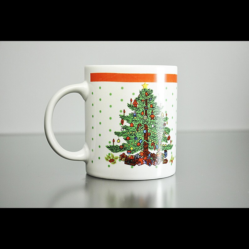 May include: White ceramic mug with a red and orange stripe around the top. The mug features a green Christmas tree with red and gold baubles, a yellow star on top, and presents at the base.