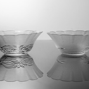 May include: Two frosted glass bowls with a scalloped rim and floral design. The bowls are reflected on a shiny surface. The bowls are likely for serving or display.