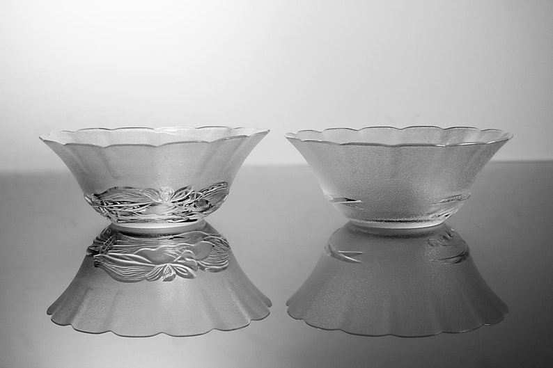 May include: Two frosted glass bowls with a scalloped rim and floral design. The bowls are reflected on a shiny surface. The bowls are likely for serving or display.