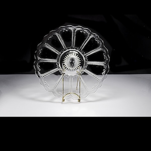 Puede incluir: Un plato de vidrio transparente con borde festoneado y un patrón decorativo de líneas radiantes. El plato está sobre un soporte de metal dorado.