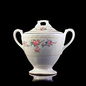 May include: White porcelain sugar bowl with a lid, featuring a floral design of pink and yellow roses with green leaves. The bowl has a gold trim around the top and base.