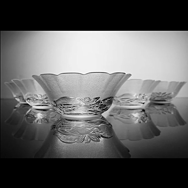 May include: A set of frosted glass bowls with a scalloped rim and floral design. The bowls are arranged in a row, reflecting on a glossy surface. The image is in black and white.