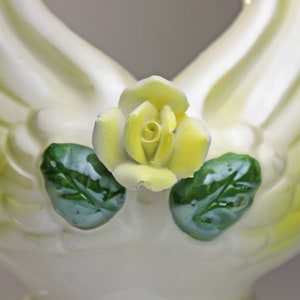 May include: A white ceramic figurine of two doves with yellow and green accents. The doves are holding a yellow rose in their beaks.