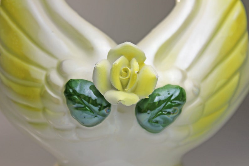 May include: A white ceramic figurine of two doves with yellow and green accents. The doves are holding a yellow rose in their beaks.