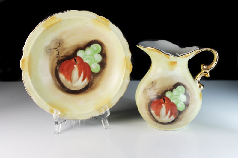 Puede incluir: Un juego vintage de vajilla de cocina de porcelana con un taz&oacute;n y una jarra de color crema con un dise&ntilde;o pintado a mano de manzanas rojas y uvas verdes.